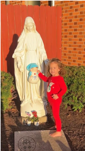 A child holding a Mary plush beside a statue of the Virgin Mary